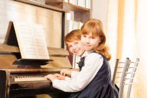 Two cute small girls in uniforms playing piano together with notes during lesson